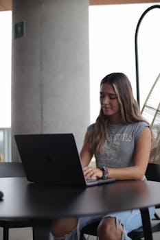 A young woman focused on her laptop, studying in a modern indoor setting.